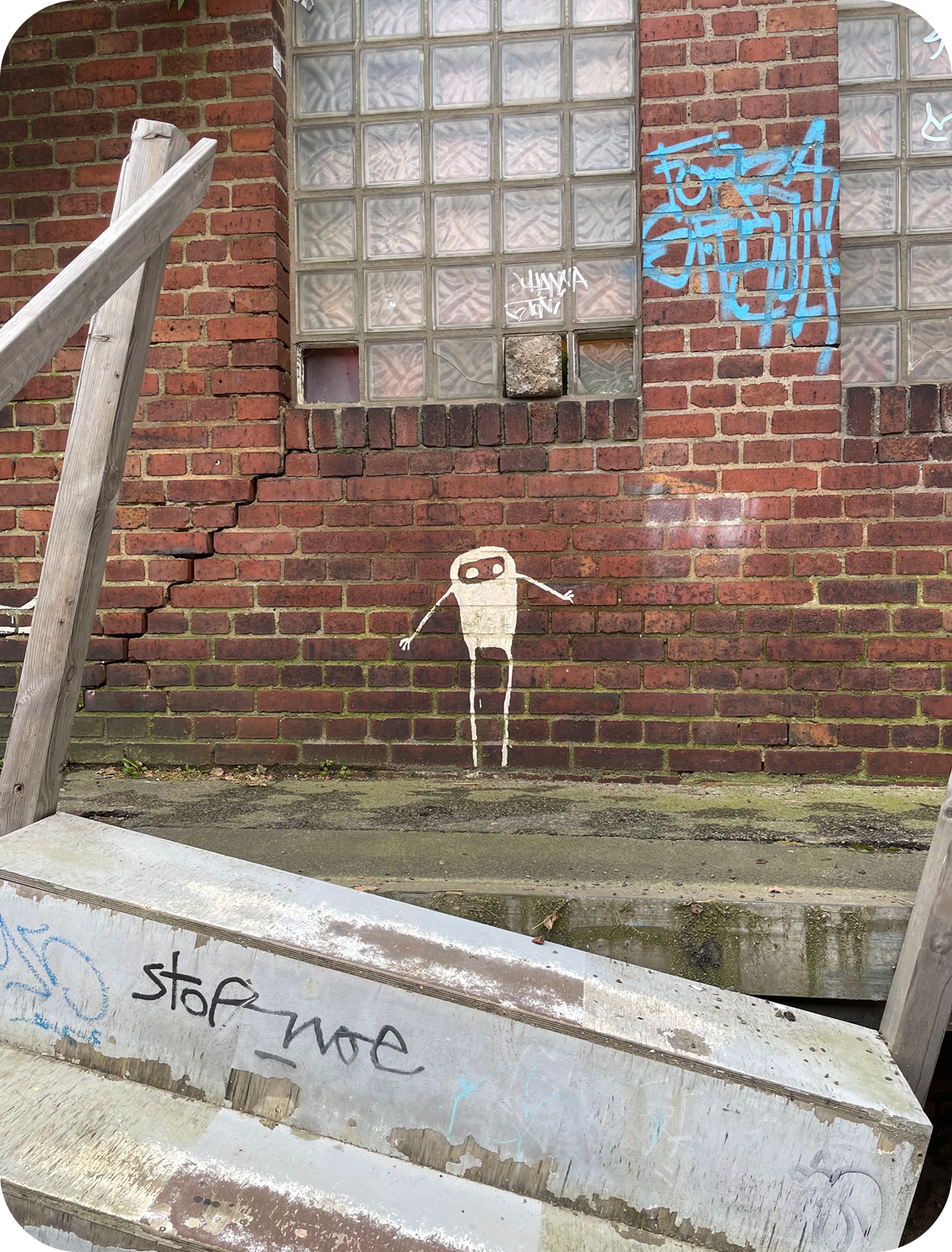 Auf einer Backsteinmauer ist eine Figur mit dünnen Armen und Beinen gemalt worden, welche ängstlich auf eine schräg stehende, alte Treppe zu schauen scheint