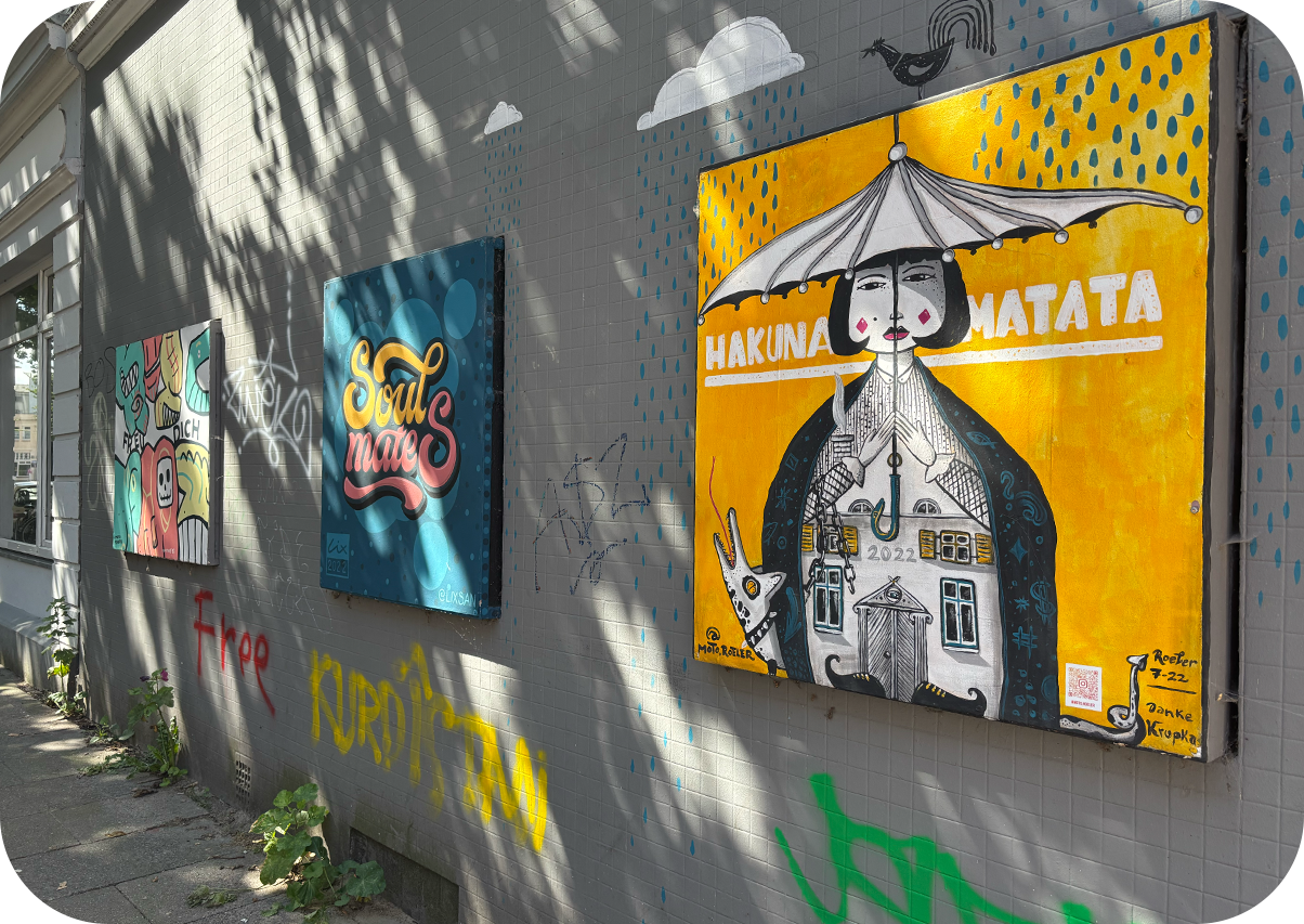 An einer Außen-Mauer an einem Wohnhaus sind drei farbenfrohe, gemalte Bilder zu sehen